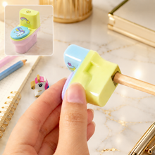 A hand holds a pastel yellow and pink toilet-shaped pencil sharpener, with a pencil being sharpened. Inset shows the full