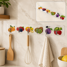 A white adhesive wall hook strip with fruit designs holds a wooden spatula, whisk, and white towel in a kitchen.