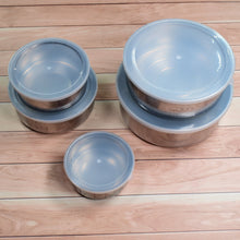 Two stacks of stainless steel food containers with clear lids, and one single container, arranged on a light wooden surface.