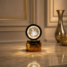 A golden base lamp with a round crystal displaying a glowing Ganesha, on a marble table with a golden vase.