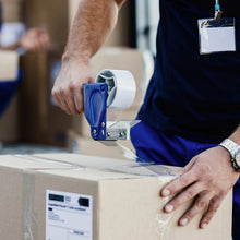 A person's hands using a blue tape dispenser to seal a cardboard box with transparent tape, showing a shipping label.