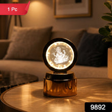 A golden lamp with a glowing Ganesha crystal sits on a round wooden table, with a blurred living room in the background.