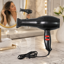 A black hair dryer with red buttons sits on a marble counter in a bathroom with a woman in a mirror reflection.