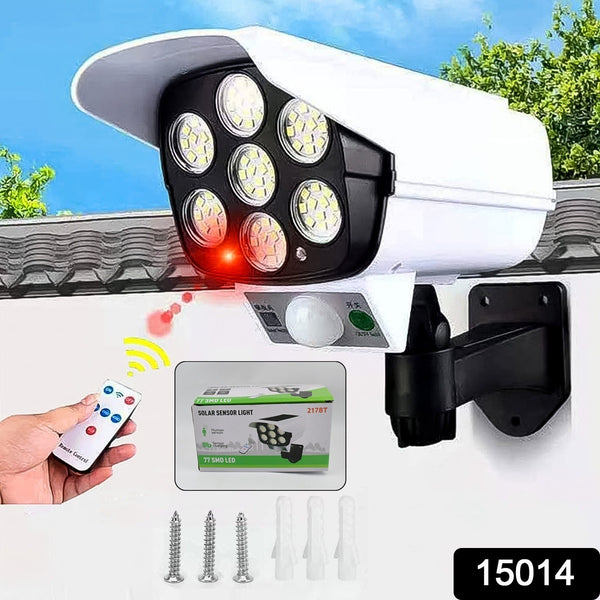 White dummy security camera with seven LED lights, a red motion sensor light, and a remote control held by a hand.