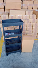 A black four-tier plastic utility trolley on wheels stands next to a brown box, surrounded by stacks of cardboard boxes in a
