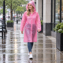 A smiling woman in a pink translucent rain coat with a hood walks on a wet city sidewalk.