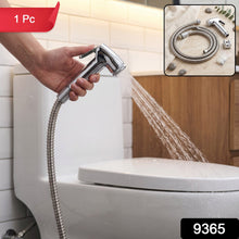 A hand holds a chrome bidet sprayer, spraying water towards a white toilet in a tiled bathroom, with a red 