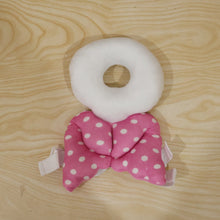A pink and white polka dot head protector with angel wings lies flat on a light wooden surface.