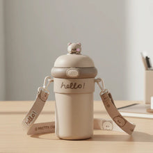 A tan sipper bottle with a bear on top, a 'hello!' inscription, and a patterned strap sits on a desk.