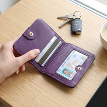 A hand holds an open purple bifold wallet with a credit card and driver's license inside, on a wooden table with keys and a