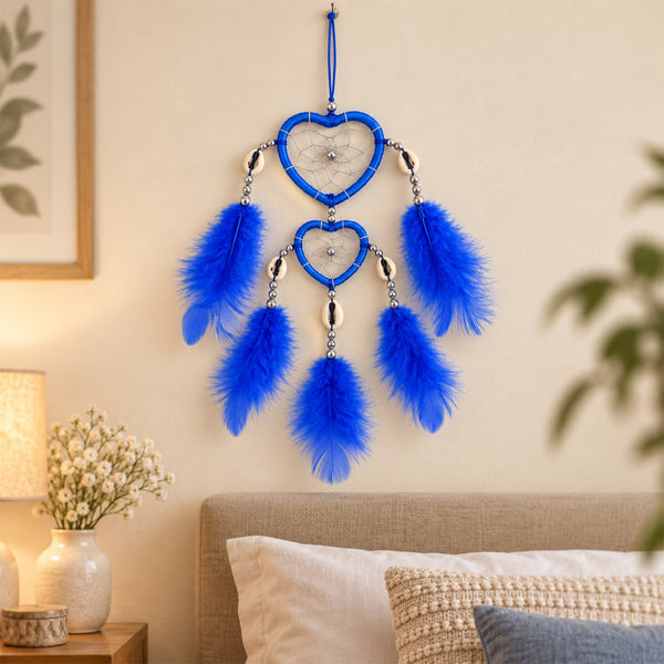 A blue heart-shaped dream catcher with feathers and silver beads hangs on a light wall above a bed.