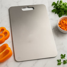 A rectangular stainless steel cutting board with a handle cutout and ruler markings on a marble surface with vegetables.