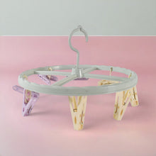 A light blue circular drying rack with purple and yellow clips is centered on a pink and grey background.