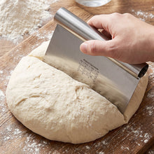 A hand uses a stainless steel dough scraper to divide a large round of dough on a wooden  board.