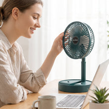 A dark green portable fan with a white USB cable on a wooden desk with a laptop and a plant.