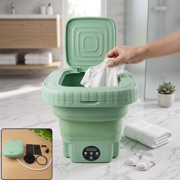 A hand places a white baby onesie into a green portable washing machine with its lid open, next to folded towels and baby
