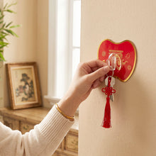 A hand hangs a set of keys with a red tassel on a red apple-shaped hook on a light-colored wall.