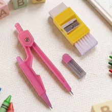 A pink compass, yellow eraser with brush, and lead refills on a white textured surface with colorful blocks.