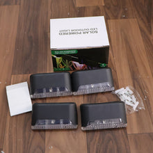 Four black solar lights, a white adhesive strip, and a bag of screws are next to an open product box on a wooden floor.