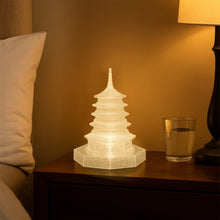 An illuminated LED crystal pagoda light on a wooden bedside table next to a glass of water and a lamp, with a white pillow.