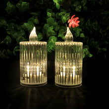 Two clear ribbed LED flameless candles with warm glow against a blurred green leafy background with a red flower.