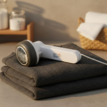 White and dark grey fabric lint remover with a USB cable resting on a stack of folded dark grey clothes.