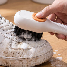 A white scrubbing brush with an orange button is actively cleaning a very dirty white sneaker, producing foam.