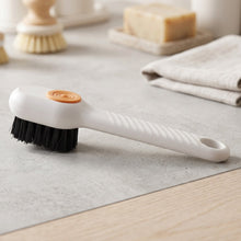 A white scrubbing brush with black bristles and an orange button rests on a gray surface with other cleaning items.