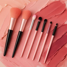 Seven makeup brushes with pink and black handles are displayed on a background with pink and red cosmetic smears.