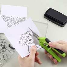 Hands use green and black snips to cut around a dog drawing on white paper, next to a butterfly drawing.