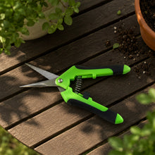 Green and black snips rest open on a wooden table next to potted plants and scattered soil.