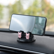 A black phone holder with two pink pig figurines on a car dashboard, holding a smartphone 