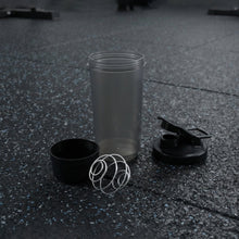 A disassembled grey and black shaker bottle with a metal whisk is on a dark gym floor.