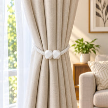 A beige curtain held back by a white braided rope tieback with geometric white plastic fasteners, in a bright room.