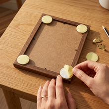 Hands applying a round beige adhesive pad to the corner of a wooden picture frame on a table.
