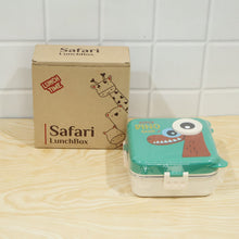 A green dinosaur lunchbox, still in its plastic wrap, sits next to a brown cardboard box with 'Safari LunchBox' branding on