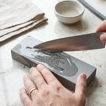Hands sharpening a silver knife on a wet gray stone with a tiger logo, next to a small bowl of water.