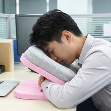 A man with dark hair sleeps on a pink desktop nap pillow with a gray cushion at an office desk.