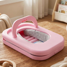 The pink desktop nap pillow is shown folded on a wooden table in a room with a bookshelf in the background.