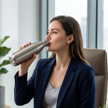 A woman in a navy blazer drinks from a silver stainless steel water bottle at her office desk, with a plant in the