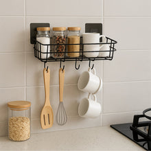 A single black metal wall-mounted storage basket with hooks, used in a kitchen to hold spice jars and hanging mugs and