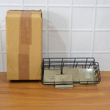 A brown cardboard box and a black metal wire storage basket with adhesive pads on a wooden surface against a white tiled