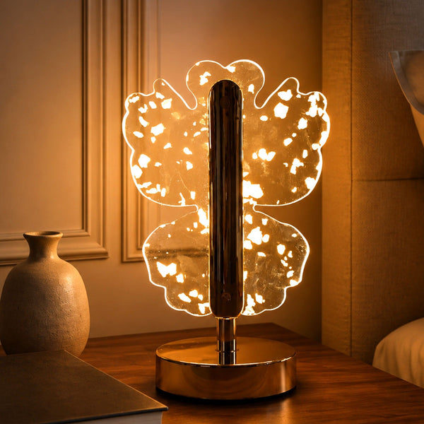 A golden butterfly-shaped LED lamp with warm light on a wooden nightstand next to a vase.