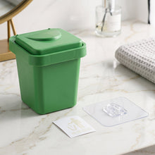 A closed green mini trash can ashtray on a marble surface with an adhesive wall mount and a small sticker.
