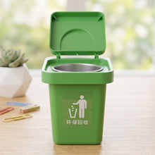 A green mini trash can ashtray with an open lid, featuring a recycling symbol, sitting on a wooden desk with a blurred