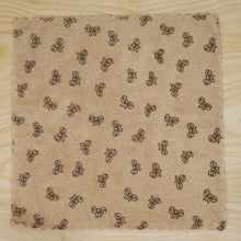A beige microfiber towel with a dark bow pattern and scalloped edges lies flat on a light wood surface.