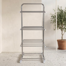 An empty stainless steel 3-layer clothes drying rack with wheels, in a bright room with a potted olive tree visible through