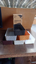 Six rectangular plastic baskets in various colors arranged on a wooden surface with a cardboard box in the background.