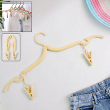 A beige foldable clothes hanger with two clips lies on a white surface next to blue jeans and a hat
